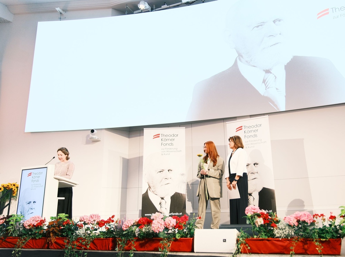 Auf einer B&uuml;hne stehen drei Frauen, eine h&auml;lt eine Rede, w&auml;hrend die anderen beiden zu ihr hin&uuml;ber blicken. &copy; TKF / Christopher Glanzl
